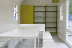 Furnishing example with green cabinet and shelf mounted in front as well as a seating area with corner bench, stools, and tables in white. PVC flooring. The tilt and turn window extending to the floor can be used as an emergency exit.