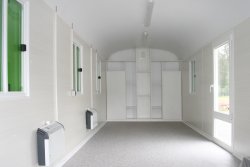 Furnishing example with white combination cabinet/shelf wall unit mounted on end, PVC flooring, plastic sliding windows, and electric wall-mounted convection heaters. The tilt and turn window extending to the floor can be used as an emergency exit.